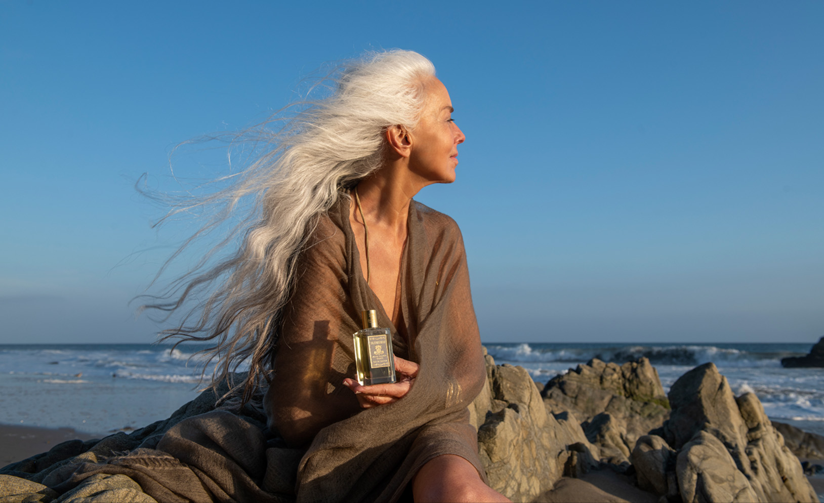 Image of Yazemeenah Rossi holding a bottle of her perfume sitting on rocks on the beach